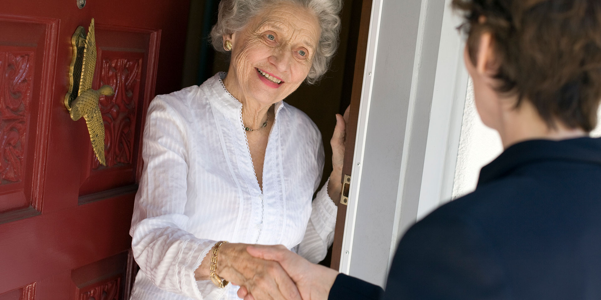 Das Bild zeigt eine alte Dame, die von einer jüngeren Dame besucht wird, beide Frauen geben sich an der Haustür die Hand Eine junge Frau besucht eine alte Dame