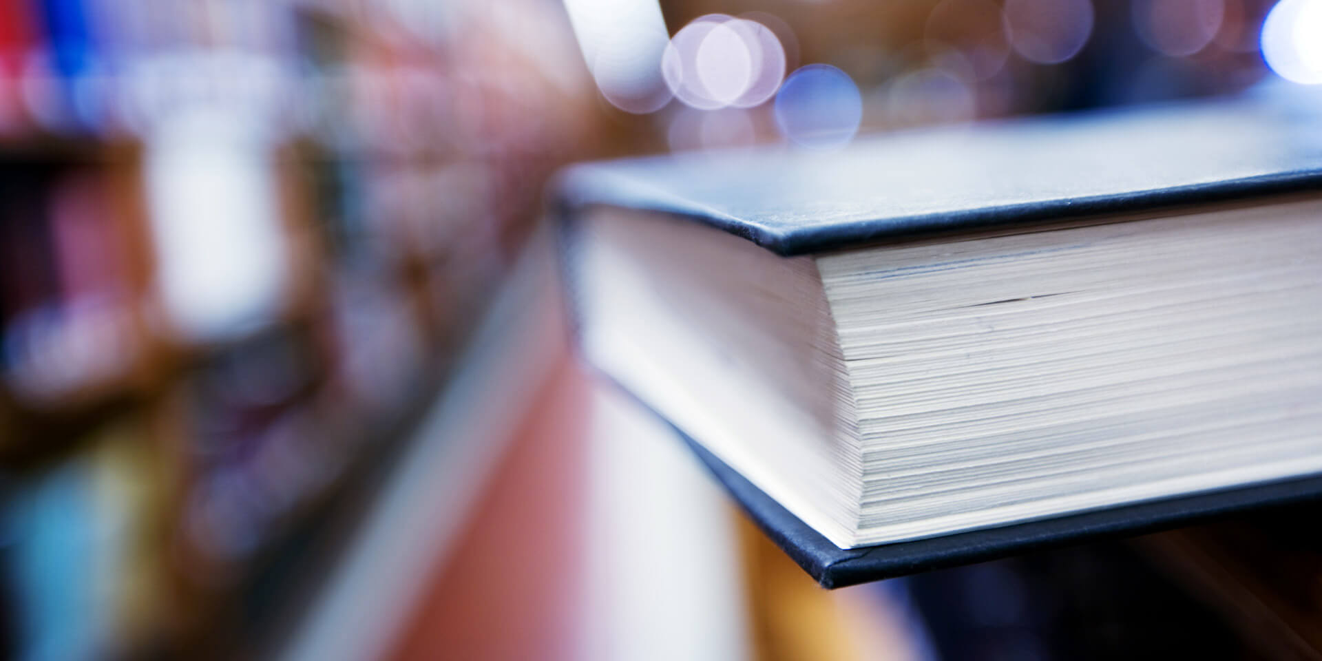 Das Bild zeigt ein Buch in einer Großaufnahme, im Hintergrund ist eine Bibliothek zu erahnen. Bibliothek