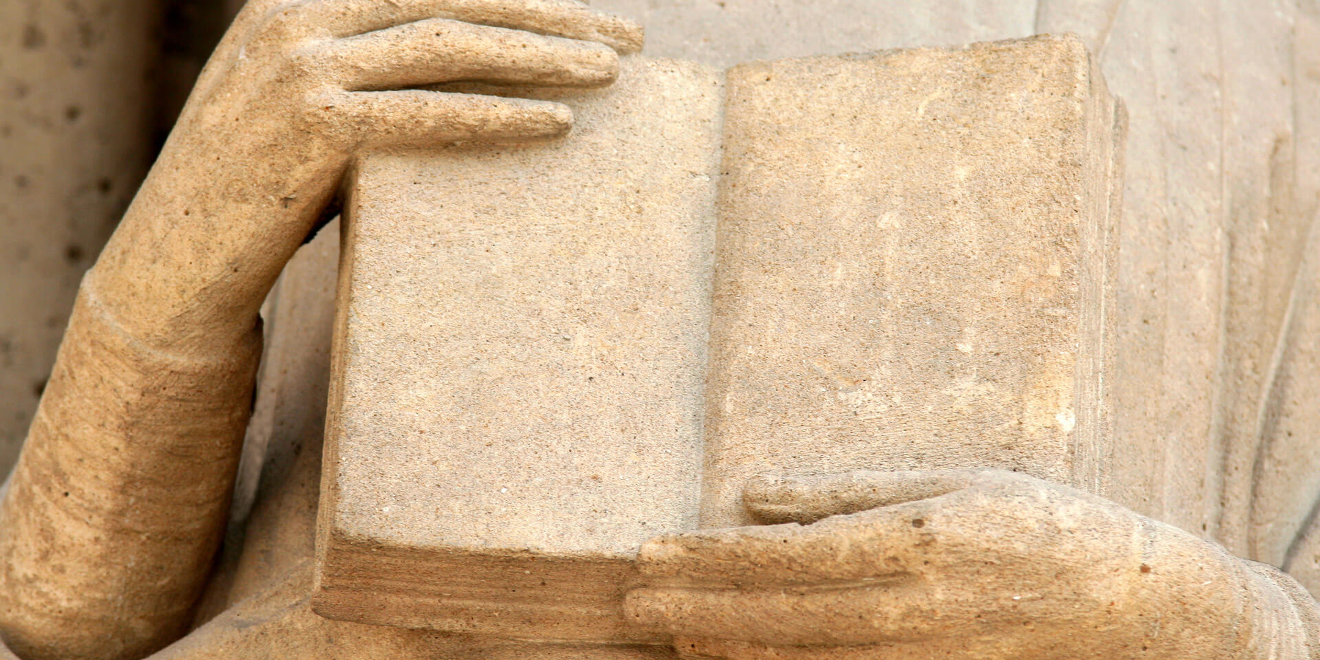 Das Bild zeigt in Großaufnahme die Hände einer Statue aus Stein, die ein Buch halten Statue aus Stein